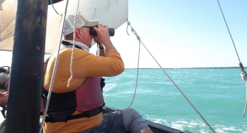 A person wearing a lifejacket sits on a sailboat and uses binoculars to look out over the blue water